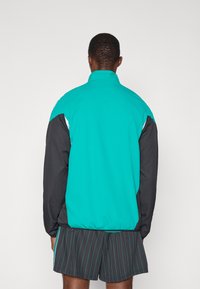 Vindjakke med en teal overdel, sorte ærmer og hvide detaljer. Fremstillet af et glat, let materiale.