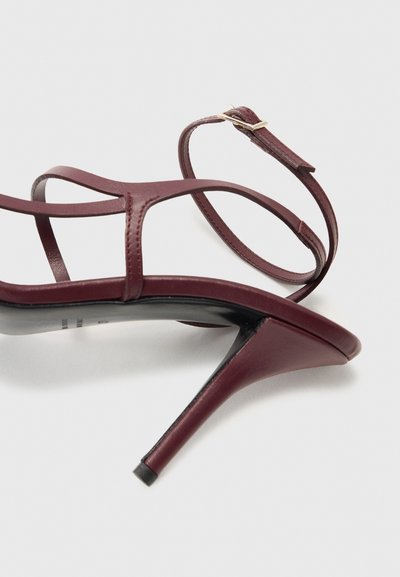 Burgundské sandály na vysokém podpatku se štíhlými pásky, přezkou kolem kotníku a strukturovaným koženým povrchem. Podpatky jsou zúžené a černé na základně.