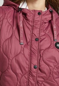 Bordeaux farbige, gesteppte Jacke mit Kapuze. Verfügt über schwarze Knöpfe und markierte Kordeln. Texturiertes Muster mit abgerundeten Formen überall.