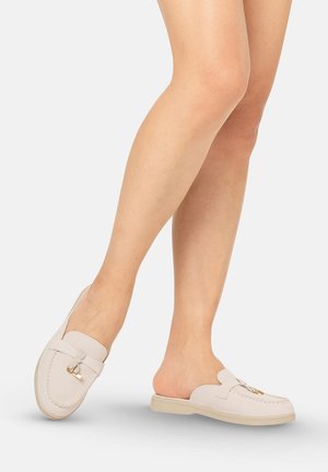 Piernas con mocasines beige tipo slip-on con herrajes dorados, sobre un fondo liso gris claro.