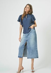 Mornarska bluza z napihnjenimi rokavi in vezanjem okoli vratu; svetlo modra denim midi krila s sprednjim razporkom; obuta v bež sandale s peto.