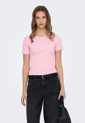 Young woman with wavy brown hair wearing a fitted pink t-shirt, black belt, black jeans, and holding a black handbag by her side.