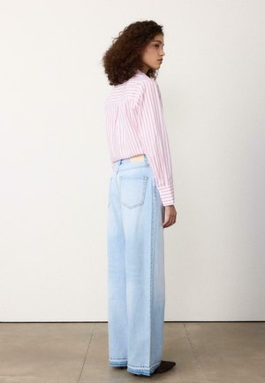 Jeune femme aux cheveux bouclés portant une chemise rose rayée rentrée dans un jean large bleu clair, debout de profil sur un sol en béton.