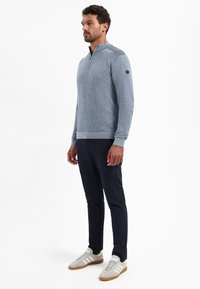 Man staat, draagt een lichtblauwe sweater met ritssluiting, donkere marineblauwe broek en witte sneakers met rubberen zolen, tegen een plain witte achtergrond.
