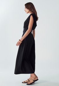 Črna obleka z prosto obliko, kratkimi rokavi, širokim pasom in teksturiranim materialom. Nosijo jo s flat sandali z metalnimi detajli.