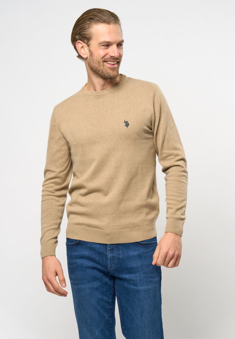 Beige trui gemaakt van zacht materiaal, met een ronde hals, lange mouwen en een klein marineblauw logo op de borst. Draagt hij met blauwe jeans.