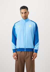 Casaco atlético azul claro com detalhes em azul escuro, possui um fecho total, gola alta, logótipo amarelo e punhos e bainha canelados.