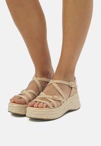 Beige wedged sandaalit, joissa on ristikkäin solmimisdesign, teksturoitu korkkipohja ja metalliset solkiyksityiskohdat. Sopii arkikäyttöön.