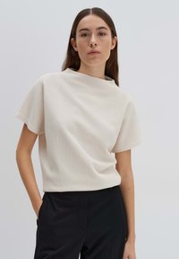 Top bianco con texture, maniche corte e collo alto, indossato con pantaloni neri. Il tessuto presenta un delicato motivo a griglia.