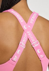 Ružová športová podprsenka s širokými priečnymi popruhmi na chrbte, bielym logom a akcentmi. Hladká textúra s nastaviteľným kovovým prvkom pre prispôsobenie veľkosti.