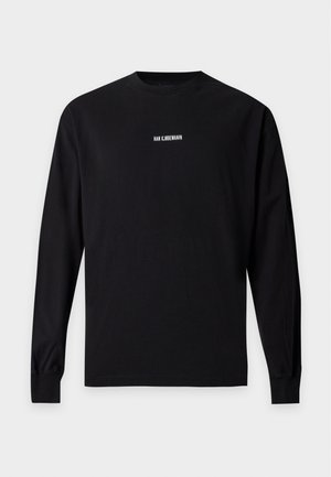 Jeune homme portant un t-shirt noir à manches longues avec un petit texte blanc, un pantalon beige large et des baskets noires, debout sur un fond uni.