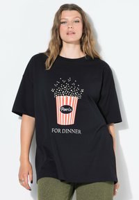 Camiseta negra de gran tamaño con un gráfico de un cubo de palomitas de maíz en rayas rojas y blancas con el texto "Perlas PARA CENAR" en blanco.