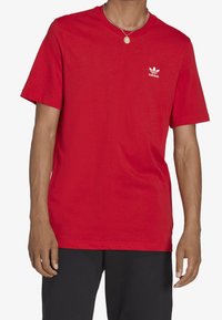 Röd bomullströja med rundad hals, korta ärmar och en vit Adidas-logotyp på vänster bröst. Stylad med svarta byxor.