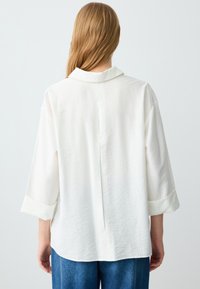 Vrouw met steil lichtbruin haar, gekleed in een losse witte langemouwen shirt en blauwe jeans, van achteren gezien tegen een witte achtergrond.