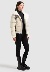 Beige puffer jacket with a plush white collar, black leather accents, and large quilted sections. Worn with black leggings and lace-up boots.
