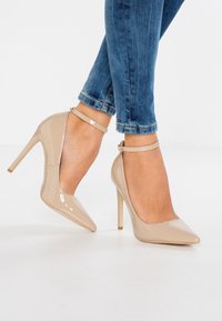 Beige lackläder högklackade skor med spetsiga tår, ankelsnodd och tunna stilettklackar. Bärs tillsammans med blå jeans som visar avsmalnande ben.