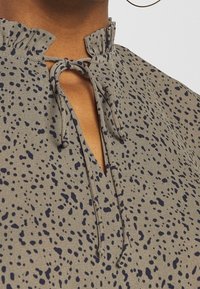 Blusa marrone con un design a motivo caratterizzato da macchie blu scuro. Il colletto ha un bordo arricciato con una chiusura a fiocco. Tessuto leggero.