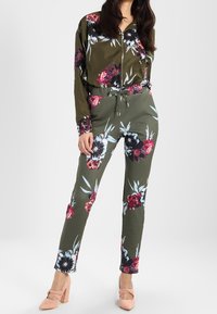 Zestaw z oliwkowozielonego materiału w kwiatowy wzór składający się z bluzki i spodni z zamkiem z przodu; ozdobiony jaskrawymi kwiatami w czerwieni i błękicie. Posiada elastyczny pasek w talii oraz zwężane nogawki.