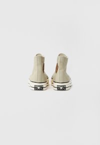 Beige High-Top-Canvas-Sneaker mit Gummisohle und schwarzen Akzenten, mit einem braunen Logopatch an der Ferse und strukturiertem Rücken.