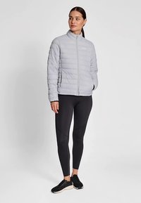 Lichtgrijze puffy jas met een ritssluiting aan de voorkant, voorzien van horizontale stiksels, gecombineerd met zwarte legging en zwarte sneakers.