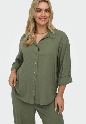 Kobieta uśmiechająca się, ubrana w oliwkowo-zieloną koszulę zapinaną na guziki z podwiniętymi rękawami oraz pasujące luźne spodnie, stojąca na jednolitym jasnym tle.