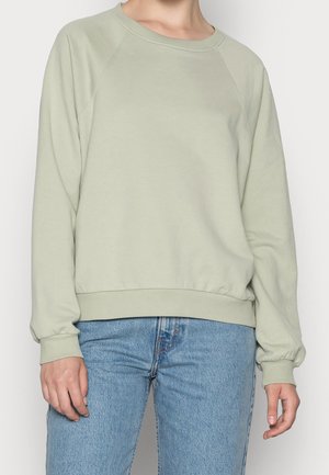 Personne portant un sweat-shirt vert clair à col rond et un jean bleu en denim, les mains détendues le long du corps.