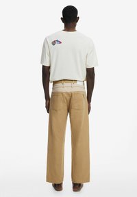 Cream short-sleeve shirt with colorful embroidered design on the back, paired with light brown wide-leg pants. Casual footwear visible.
