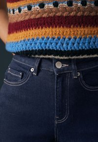 Primer plano de vaqueros denim azul oscuro con costuras blancas combinados con un jersey de punto texturizado multicolor a rayas.