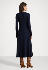 Marineblå langermet strikkekjole med tettsittende liv og svingete skjørt. Modellen har på seg brune hælsko. Glatt tekstur, enkelt design.