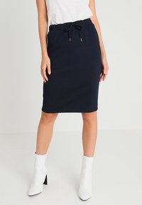 Jupe crayon en maille côtelée marine avec taille à cordon de serrage, présentant une silhouette ajustée. Portée avec un haut blanc et des bottines blanches.