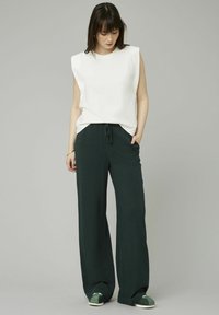 Haut sans manches blanc avec une coupe décontractée, associé à un pantalon large vert foncé avec une taille à cordon de serrage et des poches latérales. Chaussures vertes.