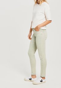 Jeans ajustados de color verde claro con una cintura alta y una textura sutil, combinados con una camisa blanca con rayas y zapatillas blancas con detalles en negro.