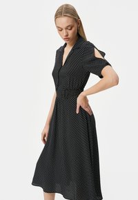Musta pilkullinen mekko lyhyillä hihoilla, napitettavalla etuosalla, vartalonmyötäisellä vyötäröllä ja hulmuavalla hameella. Kevyt kangas sileällä pinnalla.