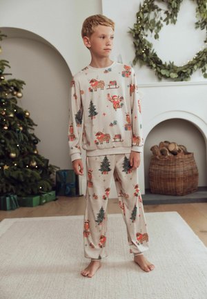 Dreng i beige julepyjamas står barfodet på et tæppe nær et pyntet træ, en pejs og en stor grøn krans i et hyggeligt rum.