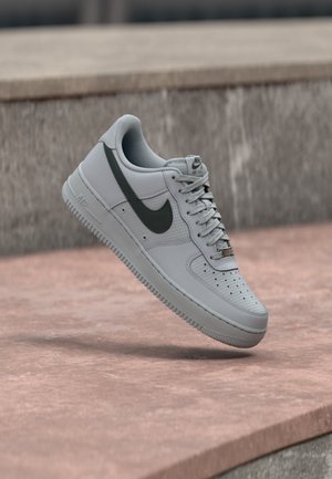 Szürke Nike Air Force 1 sportcipő fekete swoosh logóval, fehér talppal és fűzőkkel, a betonfelület fölött lebegve.