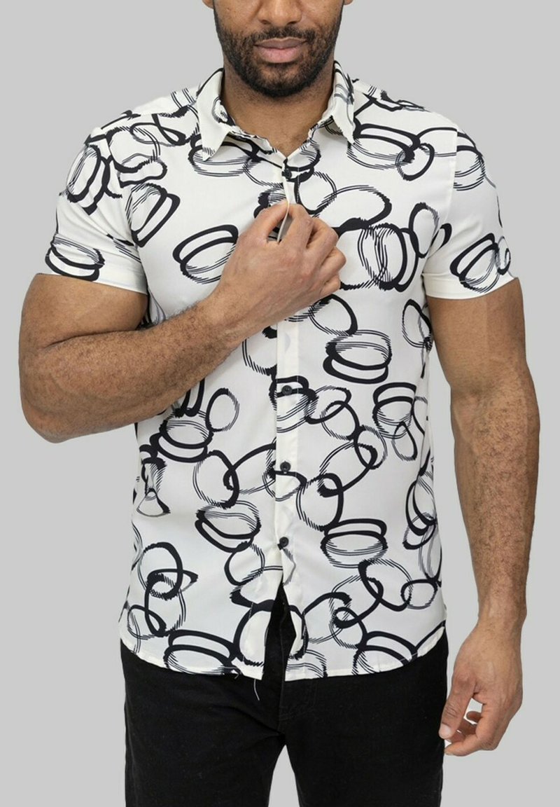 Chemise à manches courtes, à boutons, de couleur blanche avec un motif circulaire abstrait noir ; tissu lisse, col, et boutons noirs. Jean noir porté.