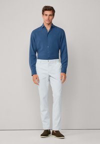 Uomo in piedi con una camicia blu a maniche lunghe, pantaloni grigi chiari e scarpe scivolate marroni scuri su uno sfondo neutro.
