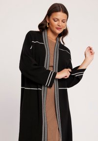 Czarny kardigan z białymi akcentami wzorowymi wzdłuż krawędzi i rękawów. Nosi się go na brązowej odzieży, ma luźny krój oraz długie rękawy.