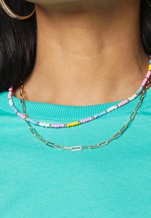 Kettingen bij een persoon die een turquoise shirt draagt: één met pastelgekleurde kralen en kleine gouden accenten, en één gouden ketting met rechthoekige schakels.