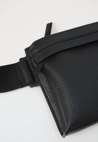 Borsa in pelle nera con texture, dotata di una zip superiore liscia, tracolla regolabile e accessori in metallo nero opaco. Design compatto e angolare con cuciture accurate.