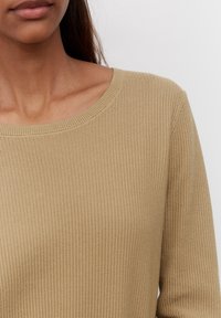 Nainen, joka on pukeutunut beigeen ribbikudottuun neulepuseroon, jossa on pyöreä pääntie, näkyy hartioista leukaan yksiväristä valkoista taustaa vasten.