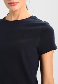 Námořnická modrá bavlněná tričko s krátkým rukávem; má malé logo v červené, bílé a modré barvě na hrudi. Měkká textura, klasický střih.