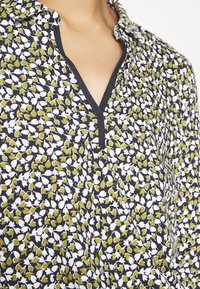 Blusa multicolore con sfondo nero e motivo floreale verde e bianco, colletto con bottoni e scollatura a V.
