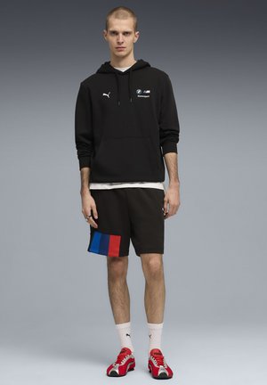 Black hooded sweatshirt with logo, black shorts featuring horizontal blue, red, and blue stripes, paired with red sneakers and white socks.