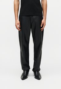 Άτομο με μαύρο δερμάτινο παντελόνι, μαύρα μποτάκια με μυτερή μύτη και μαύρο πουκάμισο, που στέκεται μπροστά σε απλό λευκό φόντο.