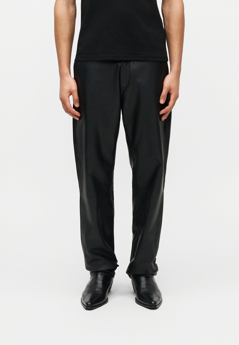 Άτομο με μαύρο δερμάτινο παντελόνι, μαύρα μποτάκια με μυτερή μύτη και μαύρο πουκάμισο, που στέκεται μπροστά σε απλό λευκό φόντο.