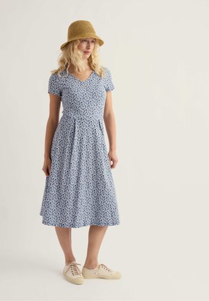 Vrouw met golvend blond haar, gekleed in een blauwe midi-jurk met bloemenmotief, beige sneakers, en een bruine gevlochten bucket hoed, staand tegen een eenvoudige achtergrond.