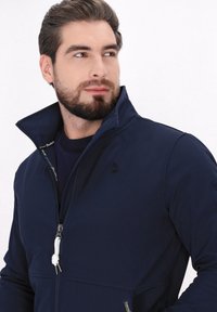 Tamsiai mėlyna zip-up striukė, pagaminta iš lygaus audinio, su aukšta apykakle, balta raišteliu ir subtiliu logotipu krūtinėje.