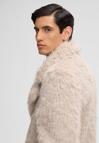 Uomo con capelli scuri pettinati all'indietro che indossa un cappotto di pelliccia sintetica beige chiaro, che guarda oltre la spalla contro uno sfondo grigio chiaro uniforme.