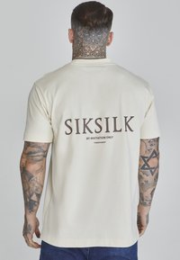 Kerman puuvillahihaton t-paita, lyhyet hihat, jossa on selässä näkyvä tummanruskea painatus "SIKSILK BY INVITATION ONLY". Yksinkertainen muotoilu, rento istuvuus.